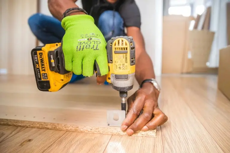 A person wearing neon green safety gloves uses a yellow power drill on a wooden surface.