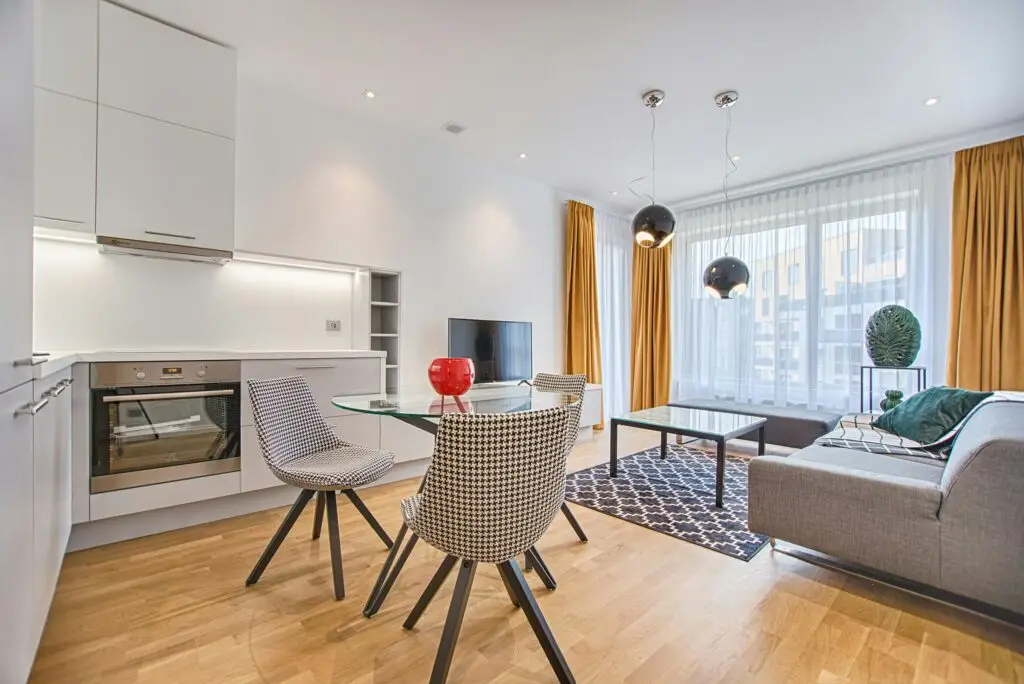 A modern studio apartment features a white kitchen, houndstooth dining chairs, yellow curtains, and a living area with a grey couch and geometric rug.