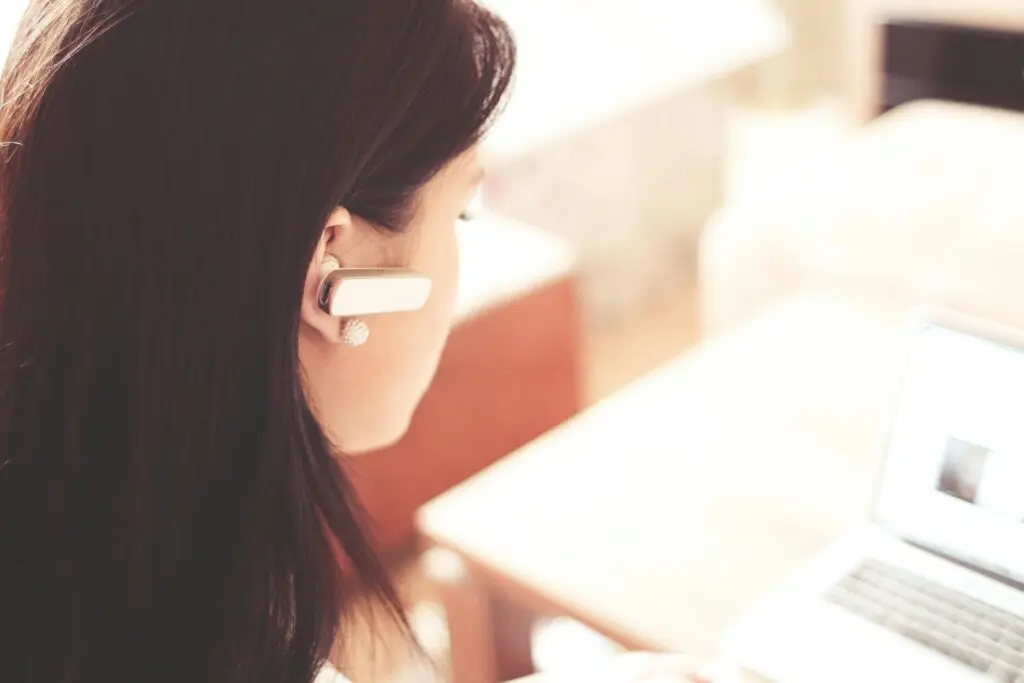 A woman wearing a Bluetooth headset works on a laptop.