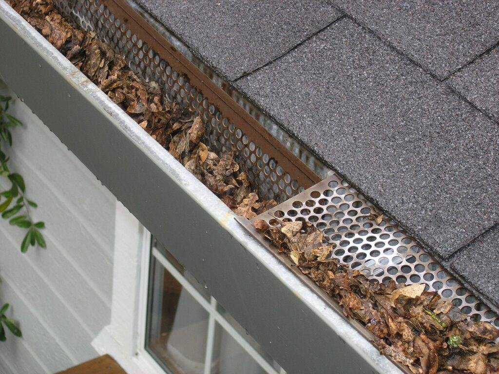 Gutter filled with leaves under a shingle roof, with a gutter guard partially visible.