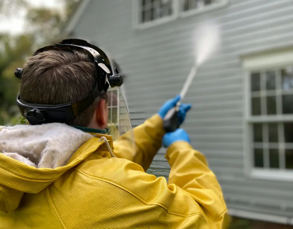 Handyman in yellow jacket and safety gear power washing a house's siding.