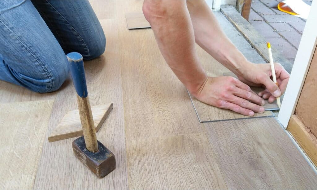 Person laying laminate flooring with hammer and pencil, measuring for the cut.